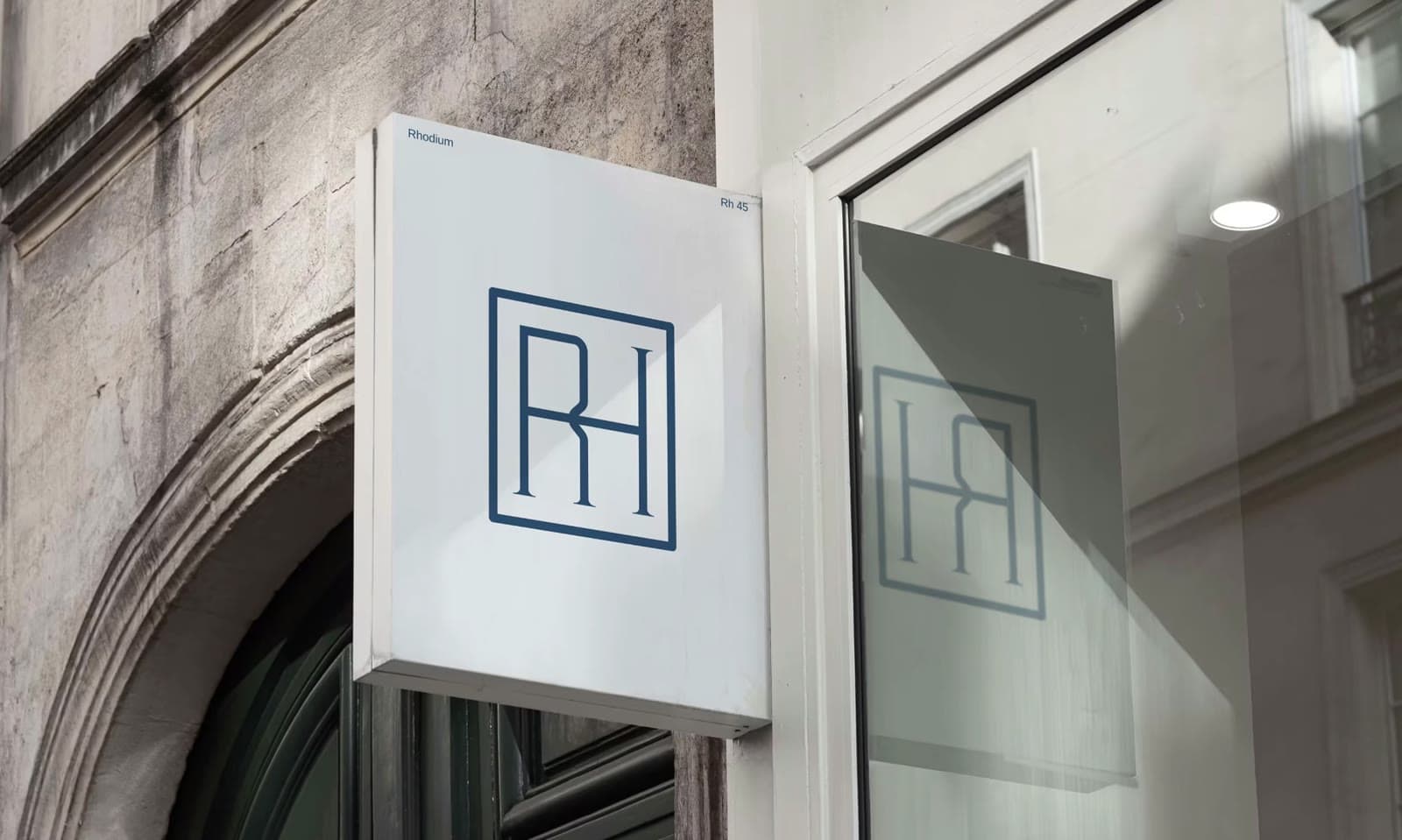 Taş cephede Rhodium tabelası: beyaz panoda lacivert çerçeveli RH monogramı ve camda yansıması